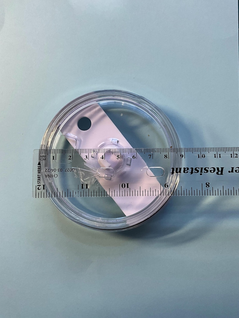 May include: A clear, circular lid with a measurement ruler laid across it. The lid has a small opening and a raised center. The ruler shows measurements in centimeters and inches. The text "Water Resistant" is visible.