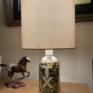 May include: A lamp made from a clear glass bottle with a white label that says "TX Blended Whiskey" and filled with small rocks. The lamp has a beige fabric lampshade.