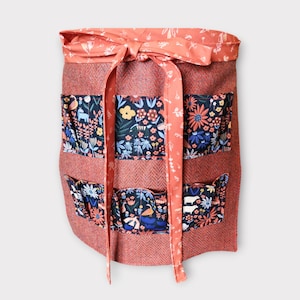 May include: A coral apron with a herringbone weave and floral pockets. The apron has a matching bow and ties. The pockets feature colorful flowers and animal designs. Ideal for gardening or crafting. The apron is designed for gardening or crafting.