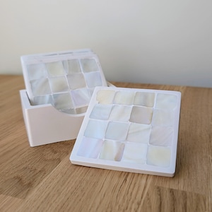 May include: A set of white square coasters with a mother-of-pearl mosaic design. The coasters are in a white holder on a wooden surface. The mosaic tiles have an iridescent sheen. The set includes multiple coasters for drinks.