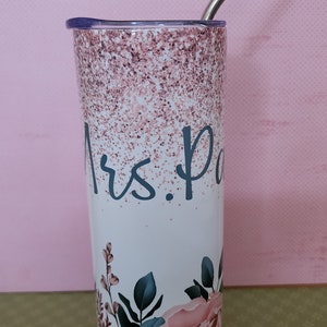 Pu&ograve; includere: Un bicchiere bianco con un design glitter rosa e un motivo floreale. Il bicchiere ha una cannuccia in acciaio inossidabile e dice "Mrs. P" sul davanti.
