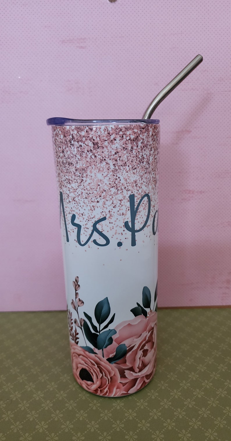 Pu&ograve; includere: Un bicchiere bianco con un design glitter rosa e un motivo floreale. Il bicchiere ha una cannuccia in acciaio inossidabile e dice "Mrs. P" sul davanti.