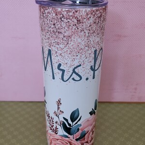 Pu&ograve; includere: Un bicchiere bianco con un design glitter rosa e un motivo floreale. Il bicchiere ha un coperchio trasparente e dice "Mrs." in scrittura blu.