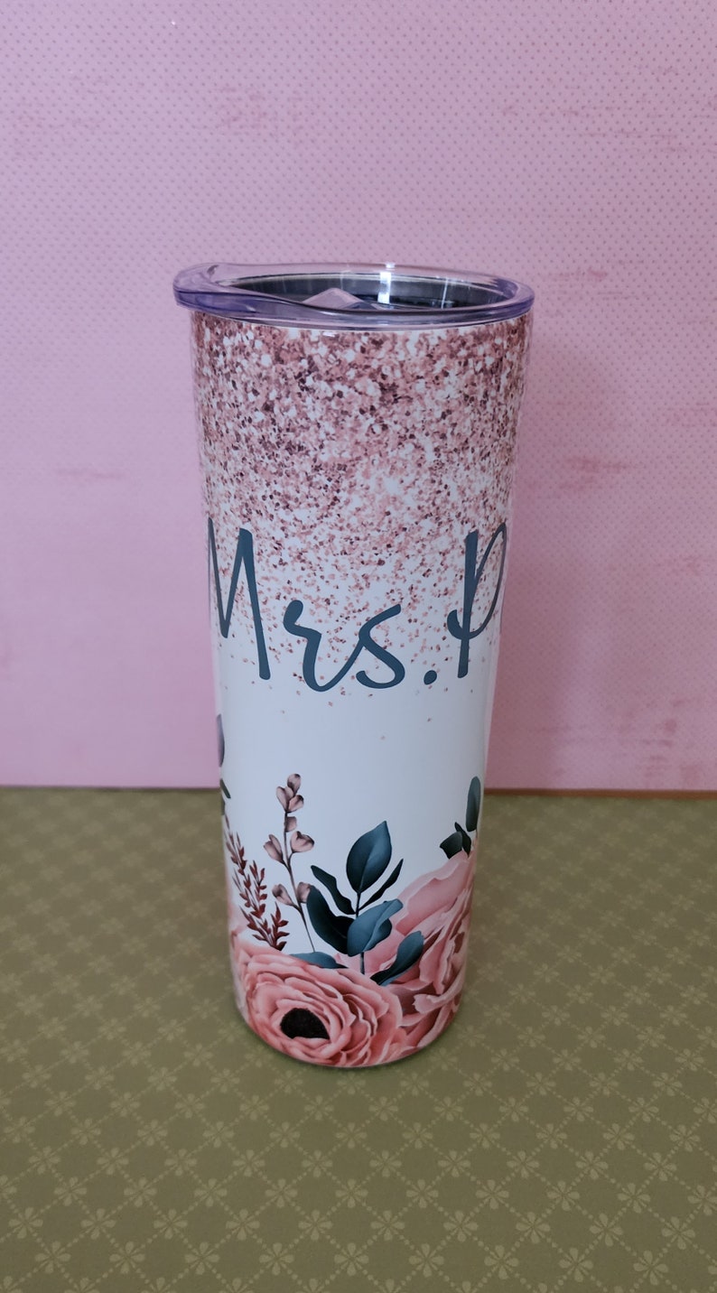 Pu&ograve; includere: Un bicchiere bianco con un design glitter rosa e un motivo floreale. Il bicchiere ha un coperchio trasparente e dice "Mrs." in scrittura blu.