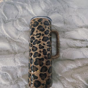 Puede incluir: Un vaso alto y aislado con asa, con un dise&ntilde;o de estampado de leopardo en manchas marrones y negras sobre un fondo dorado brillante. El vaso tiene una tapa transparente y un asa brillante a juego.