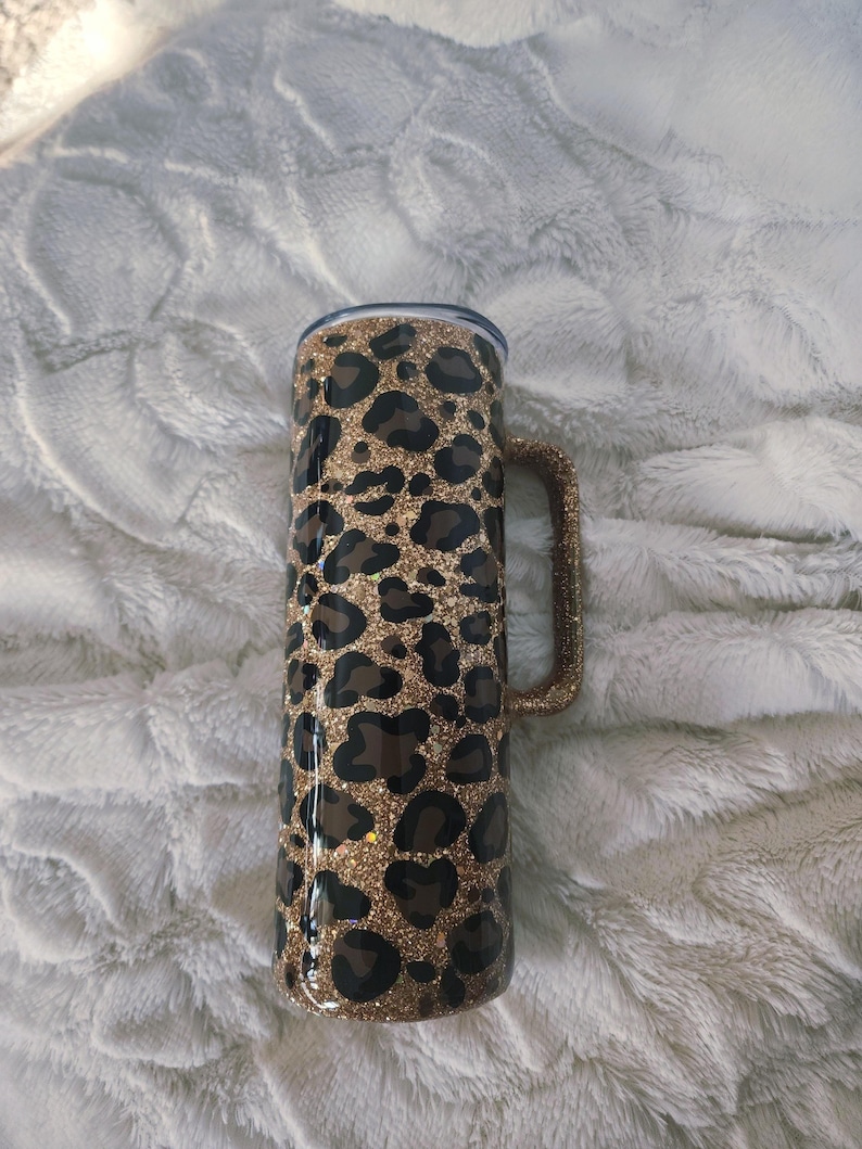 Puede incluir: Un vaso alto y aislado con asa, con un dise&ntilde;o de estampado de leopardo en manchas marrones y negras sobre un fondo dorado brillante. El vaso tiene una tapa transparente y un asa brillante a juego.