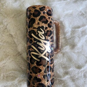 Puede incluir: Un vaso con asa y purpurina, con un dise&ntilde;o de estampado de leopardo en manchas marrones y negras sobre un fondo de purpurina dorada. El nombre "Kyla" est&aacute; escrito en cursiva en la parte delantera del vaso.