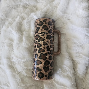 Puede incluir: Un vaso alto y aislado con asa, con un dise&ntilde;o de estampado de leopardo. El vaso es principalmente dorado con manchas marrones y un acabado brillante. El asa tambi&eacute;n es dorada y brillante.