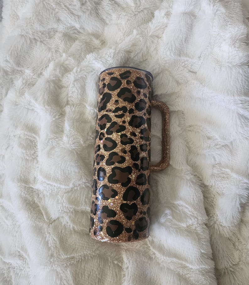 Puede incluir: Un vaso alto y aislado con asa, con un dise&ntilde;o de estampado de leopardo. El vaso es principalmente dorado con manchas marrones y un acabado brillante. El asa tambi&eacute;n es dorada y brillante.