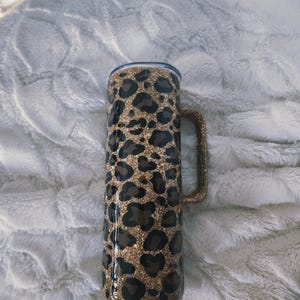 Puede incluir: Un vaso cil&iacute;ndrico con estampado de leopardo. El vaso tiene un asa y una tapa. El estampado de leopardo presenta manchas marrones y negras sobre un fondo de purpurina dorada. Ideal para bebidas.