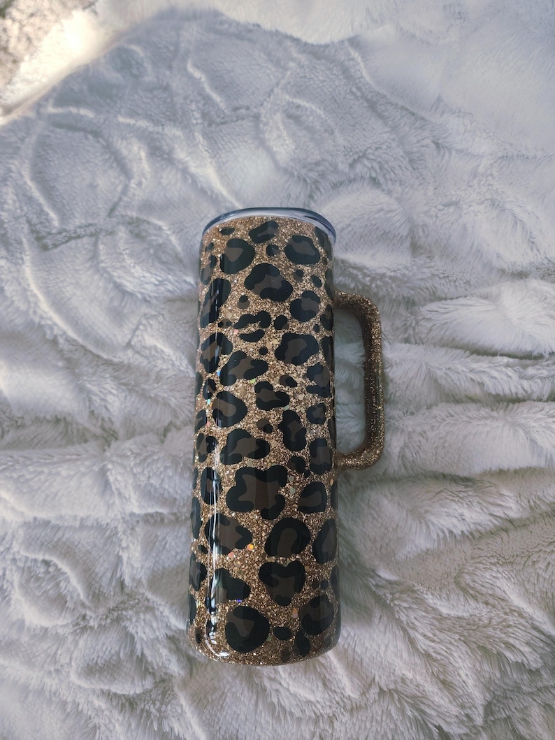 Puede incluir: Un vaso cil&iacute;ndrico con estampado de leopardo. El vaso tiene un asa y una tapa. El estampado de leopardo presenta manchas marrones y negras sobre un fondo de purpurina dorada. Ideal para bebidas.