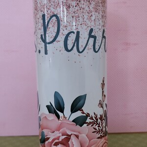 Pu&ograve; includere: Un bicchiere bianco con un design glitter rosa e un motivo floreale. Il bicchiere ha il nome "Parr" scritto in nero.