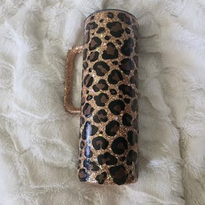 Puede incluir: Un vaso cil&iacute;ndrico con asa, decorado con un estampado de leopardo con manchas marrones y negras sobre un fondo dorado brillante. El asa tambi&eacute;n est&aacute; cubierta de purpurina dorada. El vaso es de acero inoxidable.