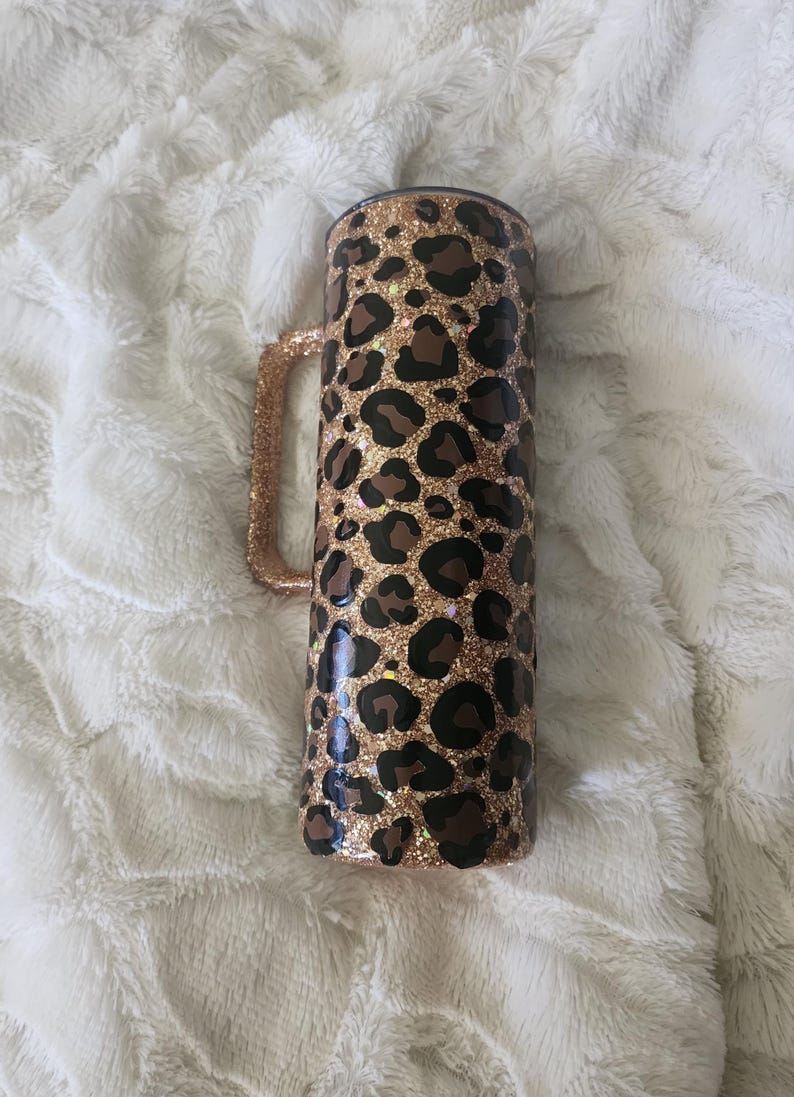 Puede incluir: Un vaso cil&iacute;ndrico con asa, decorado con un estampado de leopardo con manchas marrones y negras sobre un fondo dorado brillante. El asa tambi&eacute;n est&aacute; cubierta de purpurina dorada. El vaso es de acero inoxidable.