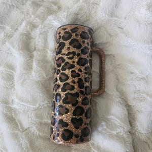 Puede incluir: Un vaso brillante con un dise&ntilde;o de estampado de leopardo. El vaso es cil&iacute;ndrico con un asa y una tapa negra. El estampado de leopardo presenta manchas marrones sobre un fondo dorado brillante. El asa tambi&eacute;n est&aacute; cubierta de purpurina dorada.