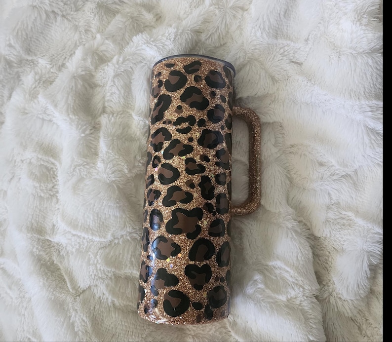 Puede incluir: Un vaso brillante con un dise&ntilde;o de estampado de leopardo. El vaso es cil&iacute;ndrico con un asa y una tapa negra. El estampado de leopardo presenta manchas marrones sobre un fondo dorado brillante. El asa tambi&eacute;n est&aacute; cubierta de purpurina dorada.
