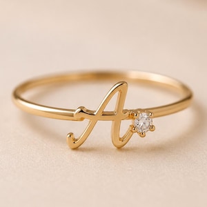 May include: A gold-toned ring featuring a cursive letter A as the central focal point. The letter is integrated into the thin, polished band and is accented by a single, small, clear round gemstone set to the right of the letter A.