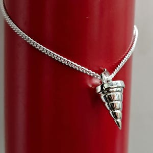 以下が含まれることがあります： 銀色のペンダントネックレス。詳細な円錐形のチャームが付いています。チャームはスパイラルデザインで、繊細なチェーンから吊り下げられています。ネックレスは鮮やかな赤色の背景に表示されています。