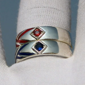May include: Two silver rings stacked together, each with a diamond-shaped gemstone. One ring has a red gemstone, the other a blue gemstone. The rings have red and blue enamel detailing.