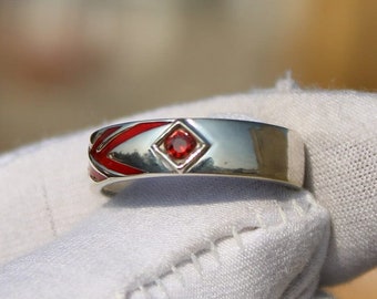 May include: A silver ring with a red enamel design and a small red gemstone in the center.