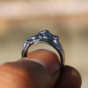 May include: A silver ring with a detailed sculpture of a reclining figure. The ring is worn on a finger.