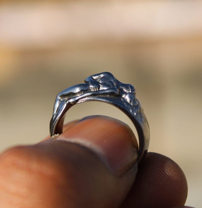 May include: A silver ring with a detailed sculpture of a reclining figure. The ring is worn on a finger.