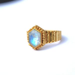 May include: A gold-colored ring with a hexagonal, iridescent gemstone. The ring has a textured band and a beaded border around the gemstone. The gemstone displays shades of blue and white, creating a luminous effect.