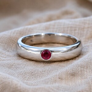 May include: A polished silver-toned band ring featuring a single, round, faceted deep red gemstone set securely in a flush bezel setting. The metal surface is smooth and reflective, showcasing a clean, rounded profile with a centered stone.