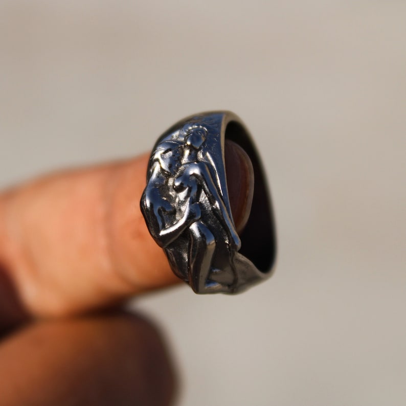 May include: A silver ring with a detailed design of two figures embracing. The ring is worn on a finger.