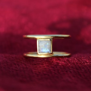 Puede incluir: Un anillo de oro con una piedra de luna de forma cuadrada engastada en el centro. El anillo tiene un diseño de doble banda.