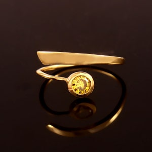 May include: A gold ring with a yellow gemstone set in a bezel setting. The ring band is a thin, twisted band that wraps around the finger.