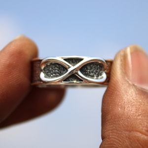 Könnte beinhalten: Ein silberner Ring mit einem Unendlichkeits-Symbol-Design. Der Ring hat ein detailliertes Muster und eine polierte Oberfläche. Das Unendlichkeits-Symbol ist der Mittelpunkt, vor einem strukturierten Hintergrund. Der Ring wird zwischen Fingern vor blauem Himmel gehalten.