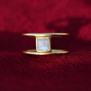 Peut inclure: Une bague en or au design à double anneau avec une pierre de lune carrée centrale. La pierre précieuse présente une qualité translucide et irisée avec de légers reflets bleus, sertie dans un cadre en métal doré lisse et poli reliant les deux anneaux.