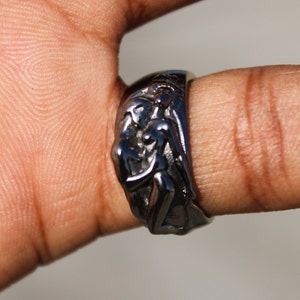 May include: A silver ring with a detailed design of two figures embracing. The ring is worn on a finger.