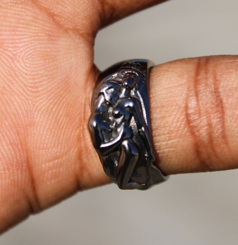 May include: A silver ring with a detailed design of two figures embracing. The ring is worn on a finger.