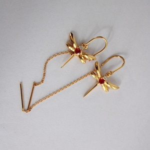 Puede incluir: Un par de pendientes dorados con forma de libélula. Cada pendiente presenta un diseño de libélula con una gema roja en el centro y una pequeña piedra transparente. Los pendientes tienen una cadena que se extiende hacia abajo con dos barras doradas en el extremo.