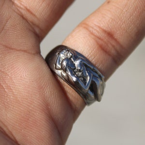 May include: A silver ring with a detailed design of two figures intertwined. The ring is worn on a finger.
