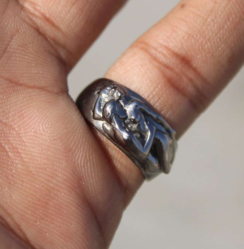May include: A silver ring with a detailed design of two figures intertwined. The ring is worn on a finger.