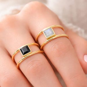 以下が含まれることがあります： 2つのゴールドリング。それぞれ正方形の宝石が特徴です。1つのリングは黒い宝石、もう1つは水色の宝石です。リングは人の指にあり、ジュエリーのデザインと色のコントラストを示しています。
