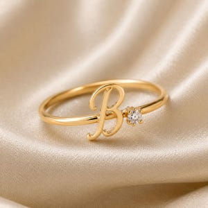 May include: A gold-toned ring featuring a cursive letter B initial as the central focal point. To the right of the letter, a single small, clear, round-cut gemstone is set into the thin, polished metal band, creating a delicate and refined appearance.