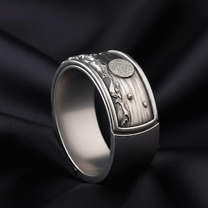 Könnte beinhalten: Ein silberner Ring mit detailliertem Landschaftsdesign. Der Ring hat ein strukturiertes Band mit einem erhabenen Design einer Berglandschaft, einem Vollmond und anderen natürlichen Elementen. Der Ring ist vor einem schwarzen Hintergrund platziert.