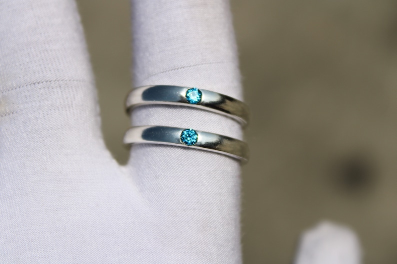 May include: Two silver wedding bands with a single blue gemstone on each band.