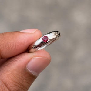 May include: A polished silver-toned metal ring featuring a single, small, round, vibrant red gemstone set flush into the center of the band. The band has a smooth, rounded profile and a reflective surface that catches the light.