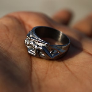 May include: A silver ring with a detailed design of two figures intertwined. The ring is made of metal and has a polished finish.