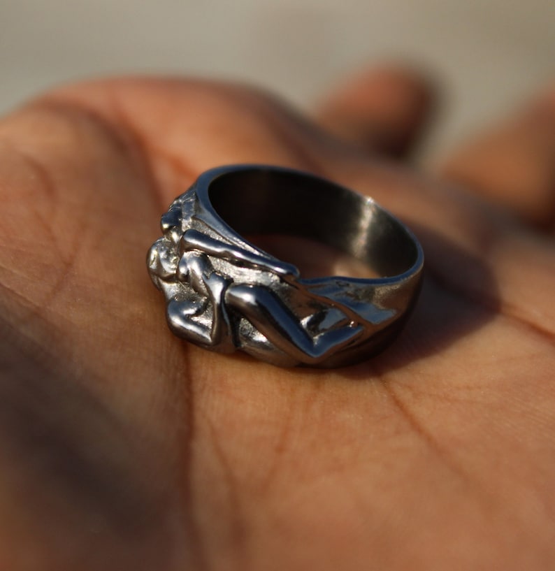 May include: A silver ring with a detailed design of two figures intertwined. The ring is made of metal and has a polished finish.