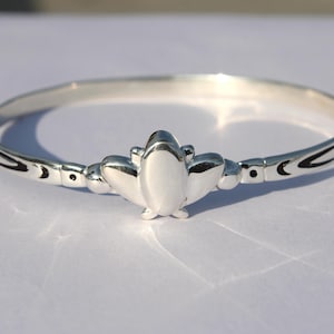Könnte beinhalten: Ein silberfarbenes Armreif-Armband mit einem zentralen dekorativen Element. Das Armband hat eine glatte, polierte Oberfläche und ein symmetrisches Design. Das zentrale Element ist eine stilisierte Form, und das Armband hat dekorative Akzente an den Seiten.