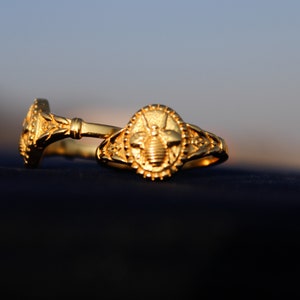 Può includere: Due anelli d'oro, uno con un designo a forma di ape e l'altro con un designo floreale, sono impilati su una superficie blu scuro.