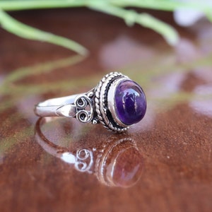 Può includere: Un anello d'argento con una pietra preziosa ametista incastonata in una montatura in argento dettagliata. L'anello presenta un design delicato e ornato con un motivo a corda intrecciata.