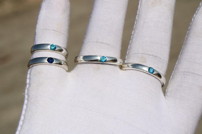 May include: Four silver rings with blue gemstones. The rings are stacked on a white surface.