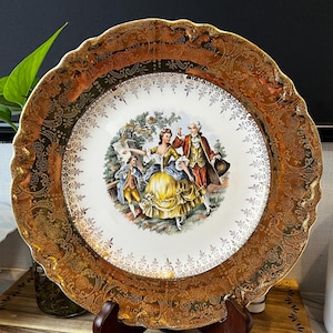 May include: A vintage porcelain plate with a gold rim and intricate gold detailing. The center features a colorful scene of a couple dancing in a garden.